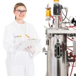 Technician taking notes beside an Infors Techfors-S 2 stainless steel bioreactor